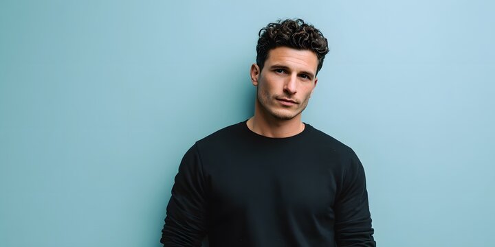 A young man with curly dark hair wearing a black shirt stands against a solid blue wall.
