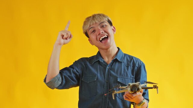 Excited young man pointing upward while holding compact drone against yellow background