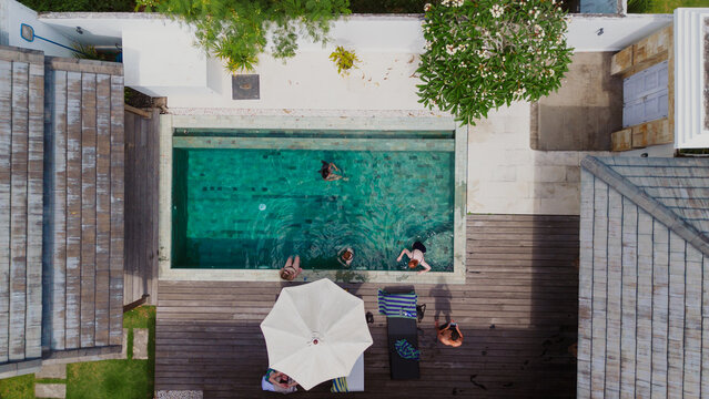 Balikpapan, Indonesia - 21 July 2026: Aerial view of people swimming in a turquoise pool at a villa with wooden decking, a white umbrella, and lush greenery.