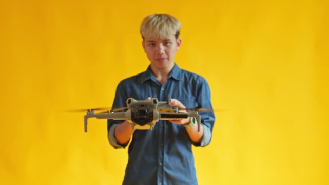 Young Asian man flying drone using remote controller against bright yellow background