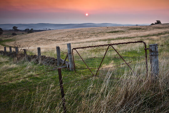 The Sun setting in a smoky sky over distant hills and a rusty farm gate