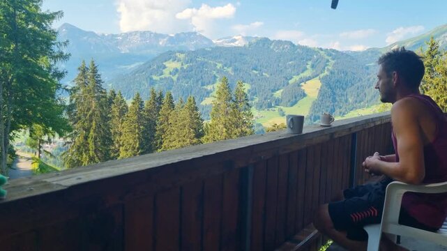 A man unwinds on the balcony of a traditional Swiss chalet in Lenk, Simmental, overlooking breathtaking Alpine scenery. The peaceful summer setting captures authentic mountain living, Switzerland