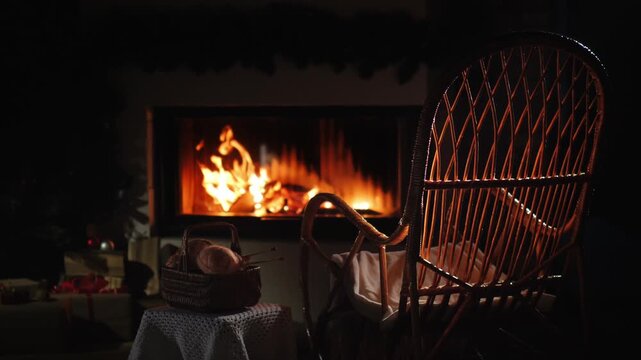 Cozy living room interior with burning fireplace, empty wicker rocking chair, and basket of yarn, offering tranquil christmas ambiance.