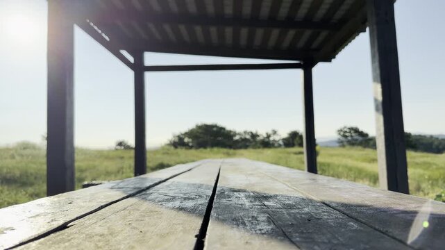 A rustic wooden table stands under an open shelter on a bright green hill. This peaceful scene brings a warm feeling of calm and quiet comfort. The sunlit field looks inviting.