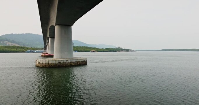 Siri Lanta Bridge spanning across water connecting Koh Lanta Noi and Koh Lanta Yai islands. Traditional fishing boats and dense coastal mangrove forest. Low angle drone flight under bridge support