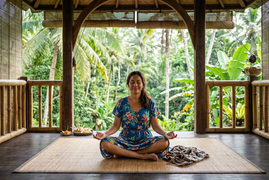 Balinese woman meditating inside an open-air yoga shala surrounded by lush tropical jungle. Mindfulness, meditation and wellness lifestyle concept