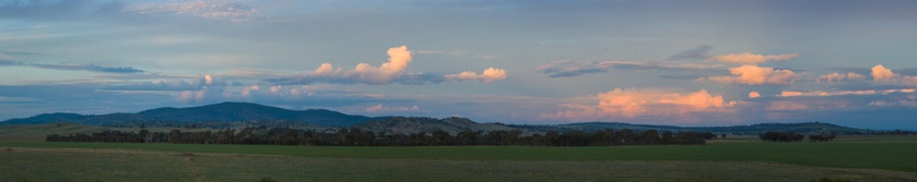 Wide panorama view of colourful clouds above hills on green farmland at twilight