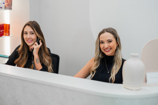 Two receptionists smiling while seated at the front desk in a skin and body clinic