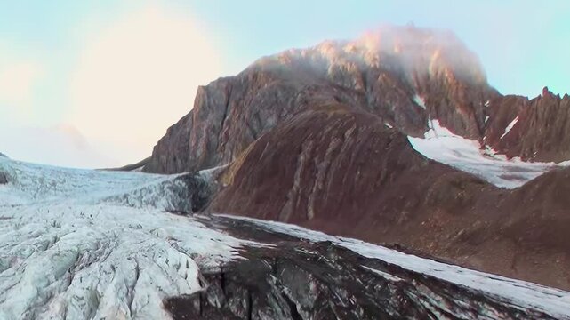 Witness breathtaking expanse of ancient glacier in pristine Arctic. Majestic ice winds its way among rugged, cloud-kissed mountain peaks under soft, golden morning sky. Serene landscape unfolds.