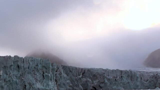Witness the colossal beauty of an ancient Arctic glacier. Its formidable icy wall stands tall against a veiled, overcast sky, reflecting the serene, stark wilderness of the polar region.