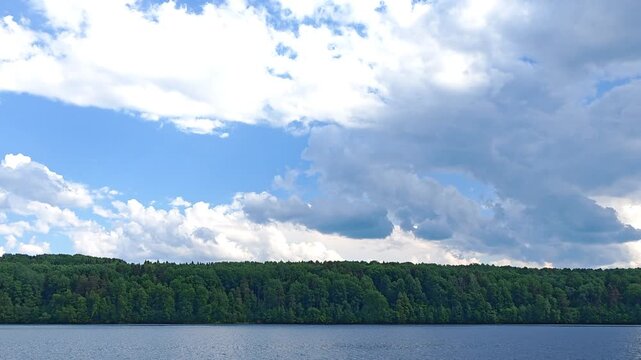 Wide panoramic view of a calm river with dense green forest on the opposite bank under blue sky with white clouds, summer nordic landscape.