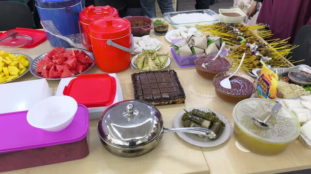 A festive Hari Raya feast features traditional Malaysian dishes on display. Served foods include compressed rice cakes, peanut sauce, and satay skewer options. Guests celebrate Eid with rich local fla