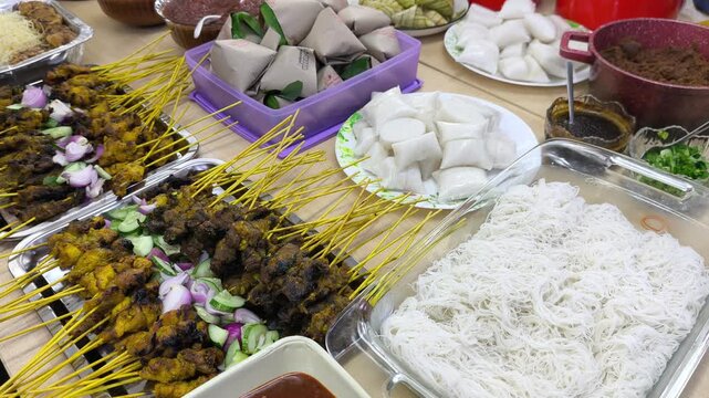 A festive Hari Raya feast features traditional Malaysian dishes on display. Served foods include compressed rice cakes, peanut sauce, and satay skewer options. Guests celebrate Eid with rich local fla