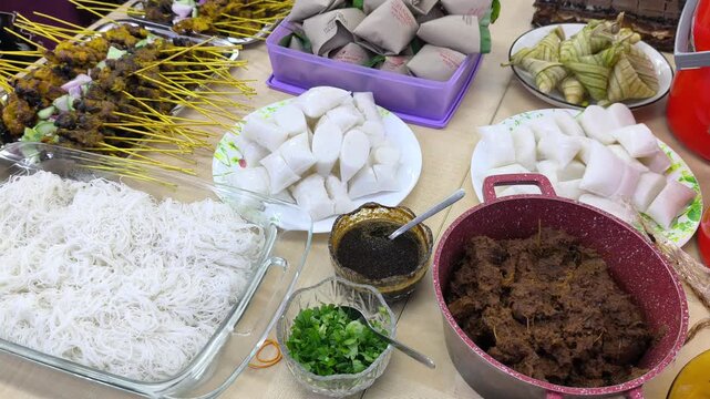 A festive Hari Raya feast features traditional Malaysian dishes on display. Served foods include compressed rice cakes, peanut sauce, and satay skewer options. Guests celebrate Eid with rich local fla