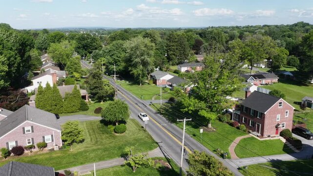 Brick colonial homes line quiet tree-shaded street in Pennsylvania suburb, spacious green lawns and mature oak trees stretch toward distant wooded countryside beyond. Aerial drone view.