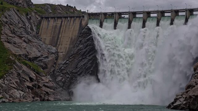 A hydroelectric dam with a dynamic waterfall viewed from the side, set in a natural environment with concrete and rocks. The scene features green and gray tones, representing clean energy.