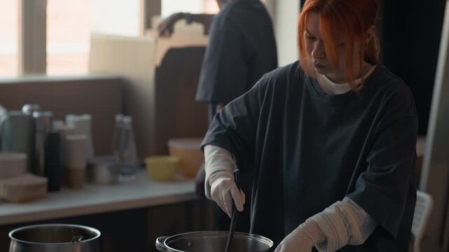 Young adult Caucasian female volunteer stirring food in large pot while diverse team packs donation boxes in background. Clip is ideal for charity campaigns and community outreach videos