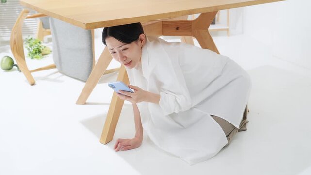 テーブルの下に潜ってスマホを見る女性。地震、緊急地震速報、アラート、防災、災害、怖い、怯える