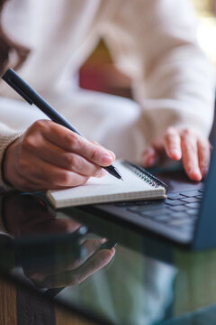 Vertical closeup image of a woman writing on a notebook while working on laptop computer