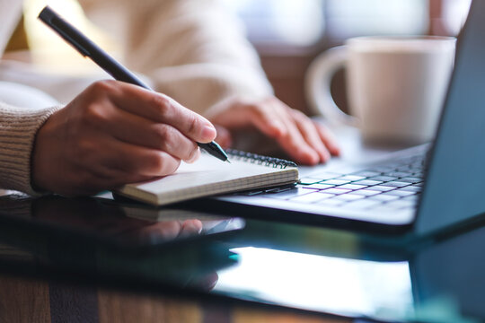 Closeup image of a woman writing on a notebook while working on laptop computer