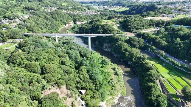 Aerial view of a river flowing through lush green hills. Modern bridge spans the river, connecting both sides. Villages dot the landscape, nestled among trees. Natural beauty surrounds the quiet