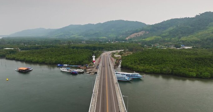 Aerial view of Siri Lanta Bridge road spanning over the water, connecting Koh Lanta Noi with the Krabi mainland, showcasing lighthouse, boats, and lush mangrove forests on tropical hills. Drone flight