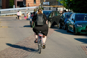 Fototapeta premium Rear view of an overweight woman in headphones riding a bicycle on a city street pavement. Plus size female cyclist commuting on a bike.