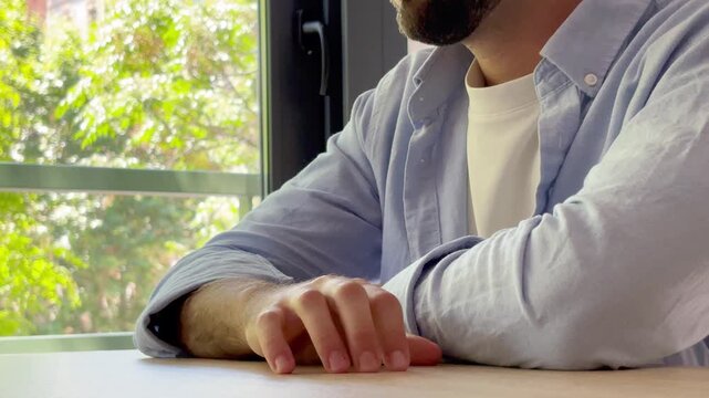 Worried and anxious young man sitting at a table next to a window, nervously tapping his fingers and looking away thoughtfully, feeling stressed and impatient while waiting for something