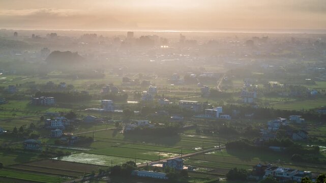 Aerial view of Lanyang Plain in Yilan, Taiwan, with green rice fields and distant city buildings covered in morning mist under a golden sunrise.