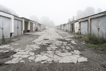 Spękana droga prowadząca przez aleję osiedlowych garaży skrytych w gęstej mgle. Mroczny, nostalgiczny klimat, urbanistyka, zniszczona nawierzchnia. © Henryk Niestrój