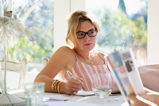 young woman wearing reading glasses taking notes at table in light filled room