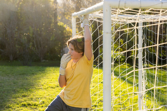 portrait of teenage boy with soccer ball framed in net at sunset
