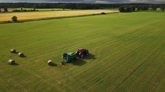 Aerial view of a tractor parked with a round baler among hay bales in a cut grass field, illustrating machinery used for forage harvesting and livestock feed production.
