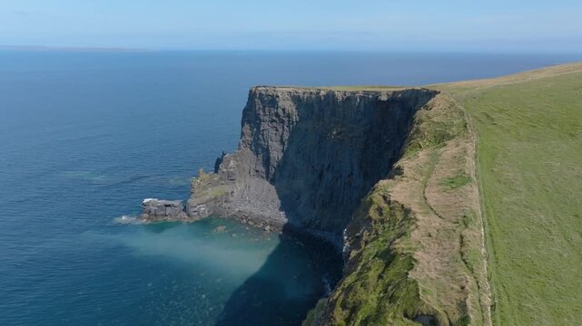 Steep coastal bluff drops from a narrow grassy path into turquoise sea below as the camera glides along the headland edge.
