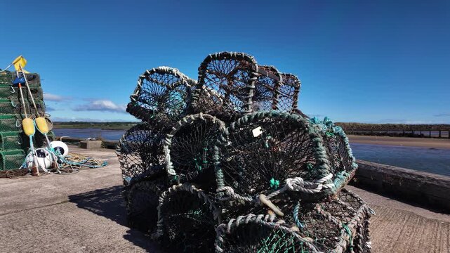 Pile of wire crab pots and coiled ropes sits on a concrete dock beside calm marina water under a clear blue sky.