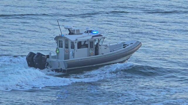A grey marine police patrol boat with flashing blue lights speeds through choppy blue ocean water on a daytime maritime security patrol.