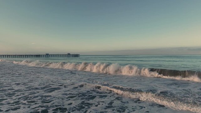 A breathtaking cinematic drone flight gliding just above the waves at sunrise, traveling along the California coast toward the iconic San Clemente Pier.
