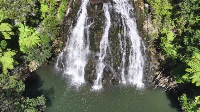 Drone footage moves closer over Owharoa Falls as multiple streams cascade down rugged rock into a serene pool, surrounded by dense rainforest in Karangahake Gorge, Waikato, New Zealand.