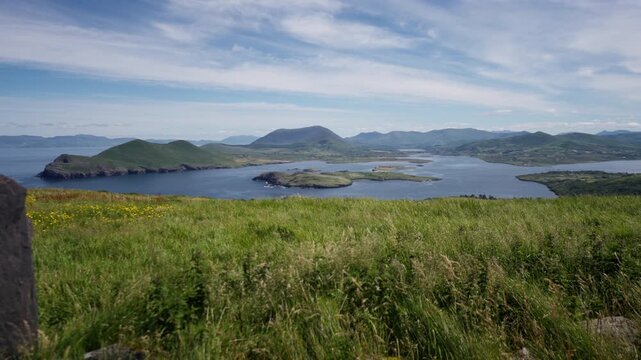 Wide view of rolling green hills and coastal islands under a blue sky with scattered clouds and distant mountain ranges.