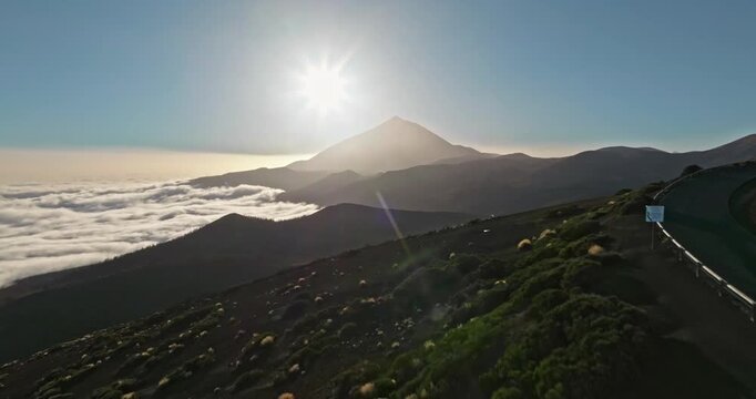 Aerial view capturing the iconic Teide volcano silhouetted against a bright sun, with a winding mountain road alongside deep valleys covered by a sea of clouds at sunset in Tenerife
