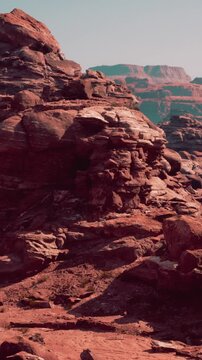 Large rock formations in a desert area. The bright sunlight highlights the texture and colors of the rocks. The landscape appears dry and rugged, with distant mountains.