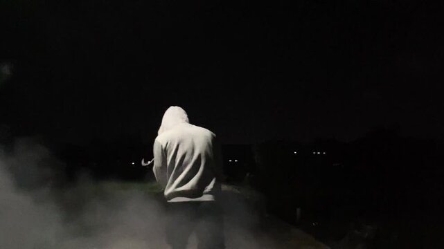 Slow motion cinematic clip of an anonymous man in a hoodie and boxing gloves practicing shadow boxing punches surrounded by thick smoke in the dark night. Intense athletic training.