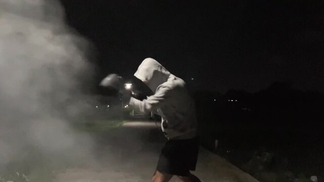 Slow motion cinematic clip of an anonymous man in a hoodie and boxing gloves practicing shadow boxing punches surrounded by thick smoke in the dark night. Intense athletic training.