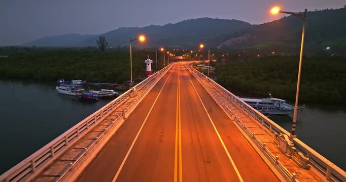 Siri Lanta Bridge at night spanning river with boats moored at the side, surrounded by mangrove forest and mountain green hills under twilight evening sky with illuminated street lights. Drone flight