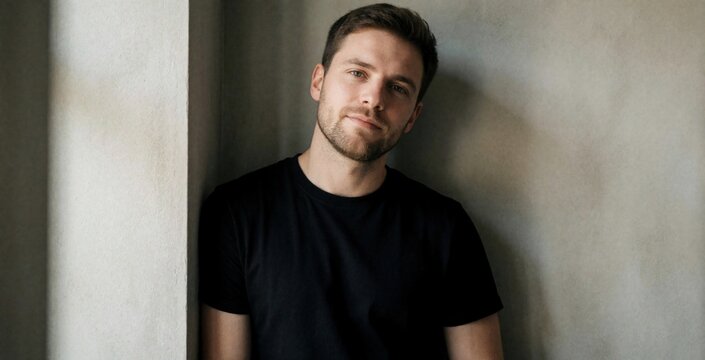 Young man in black t-shirt leans against textured wall. Casual, relaxed pose, friendly expression, looking towards camera. Natural light illuminates features, creating simple yet striking portrait.