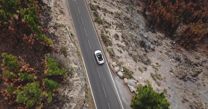 White convertible car traveling through a barren, rocky mountain landscape with patches of green pine trees and areas of dry, burnt looking vegetation on the island of Tenerife. Aerial top down view