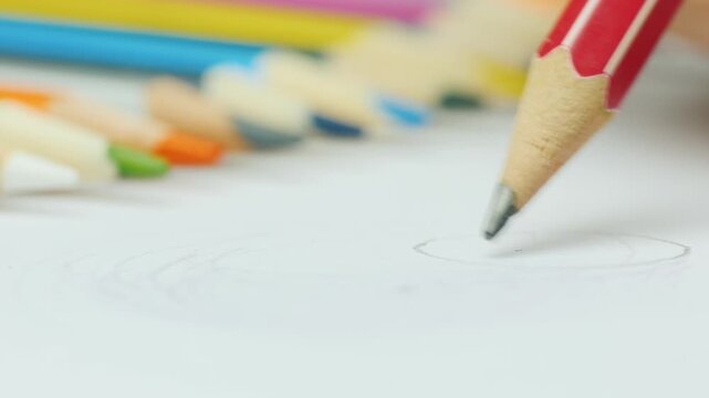 Close up of pencil drawing circle on white paper with blurred colored pencils in background.