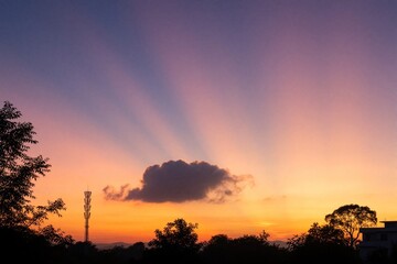 Naklejka premium Vibrant sunrise sky with purple, orange gradient colors. Golden sunbeams radiate through single dark cloud over silhouette of trees, cell tower. Morning horizon scene features natural beauty,