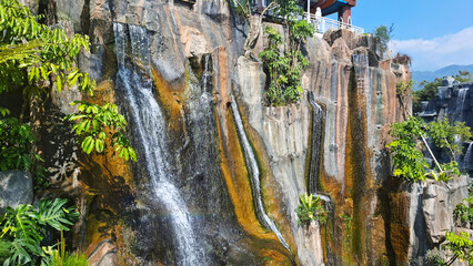 a tourist spot with a magnificent artificial waterfall and mossy cliffs surrounded by refreshing tropical vegetation under bright blue sky © umi