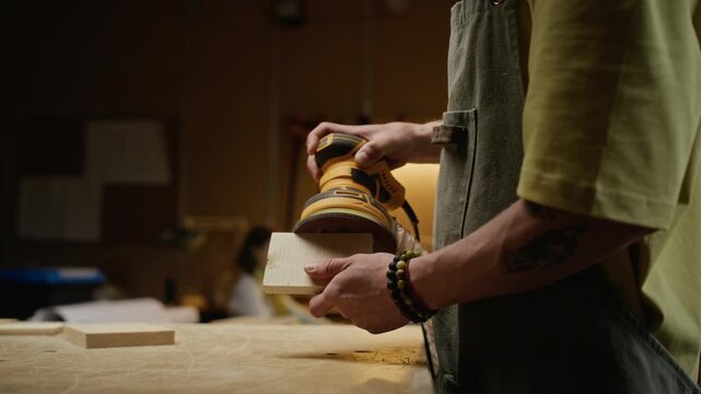 Young adult Caucasian man wearing safety glasses and apron sanding small wooden block with orbital sander in carpentry workshop. Footage is ideal for small business marketing and craftsmanship ads